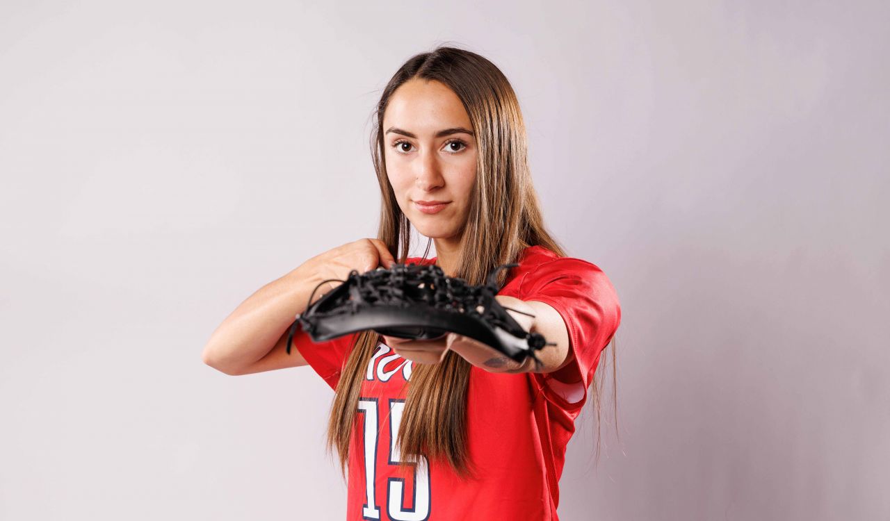 Ryanne logon posing with a lacrosse stick and wearing red Brock University varsity lacrosse jersey in front of a grey background.