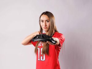 Ryanne logon posing with a lacrosse stick and wearing red Brock University varsity lacrosse jersey in front of a grey background.