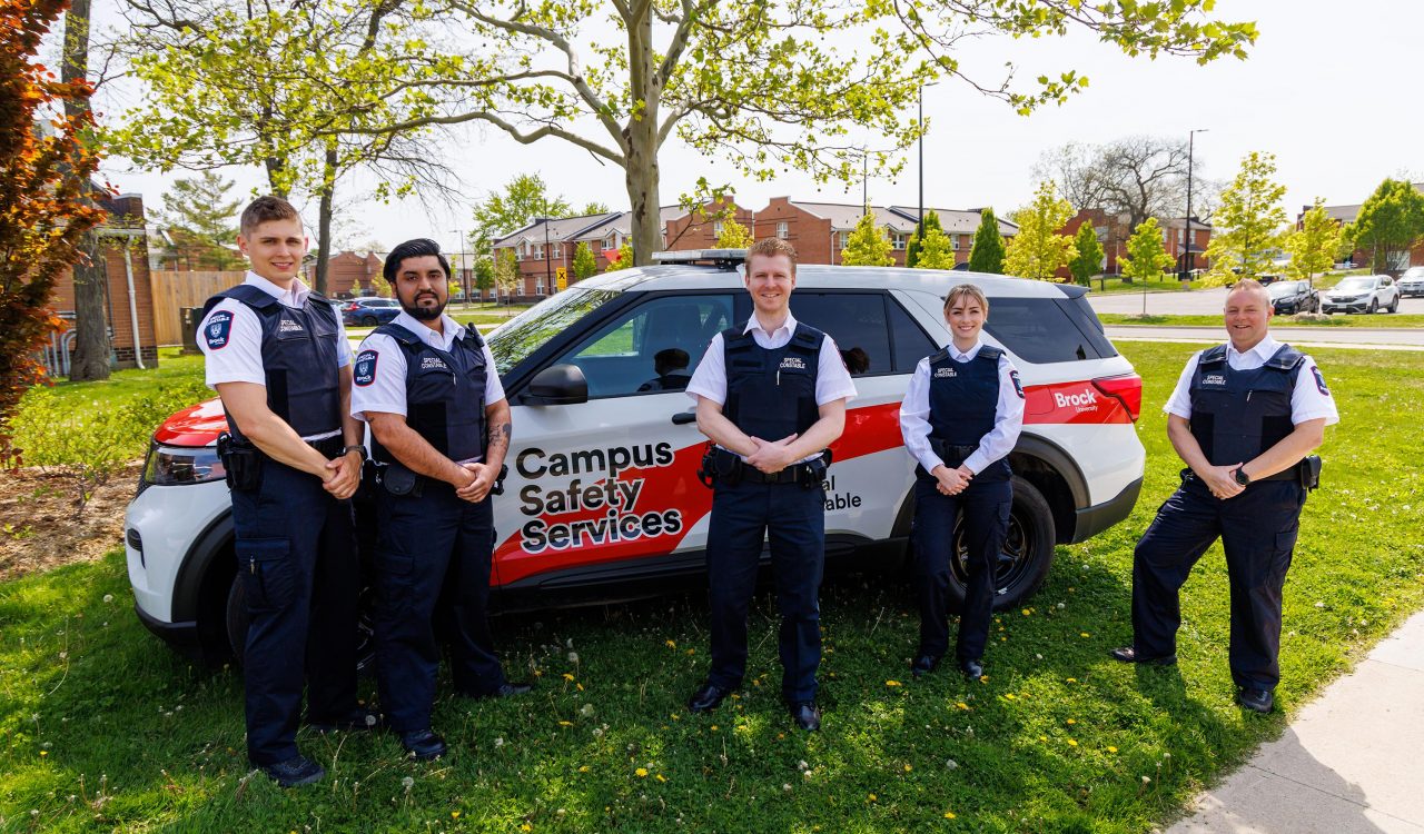 Five uniformed officers stand beside a Campus Safety Services vehicle. 