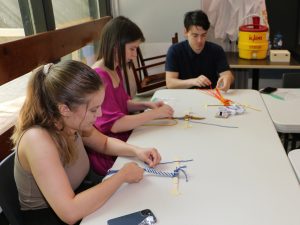 Three people sit at an indoor table weaving small sashes out of string.