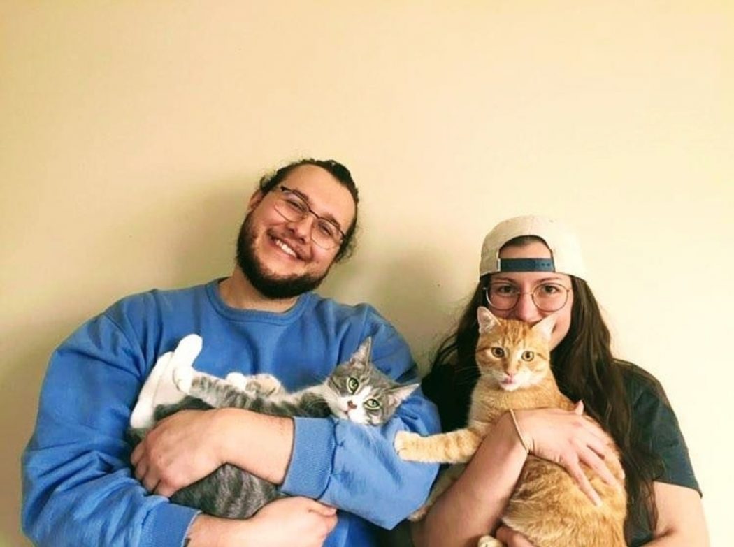Mac Orlando and Mandy Nemec-Bakk sit in front of a wall each holding a cat while smiling.