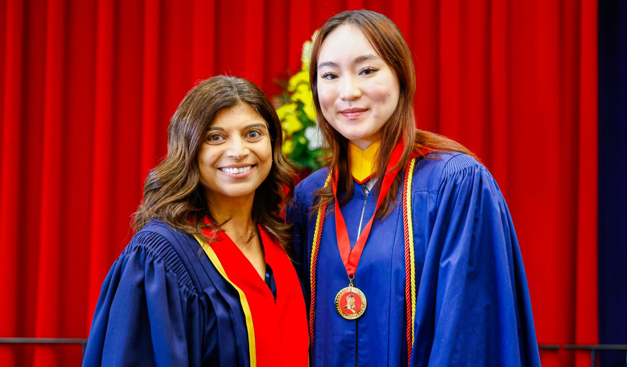 Arti Freeman (left) presents Silvana Nguyen with a Spirit of Brock medal on stage during Brock University’s Convocation ceremony. They are each wearing academic regalia.