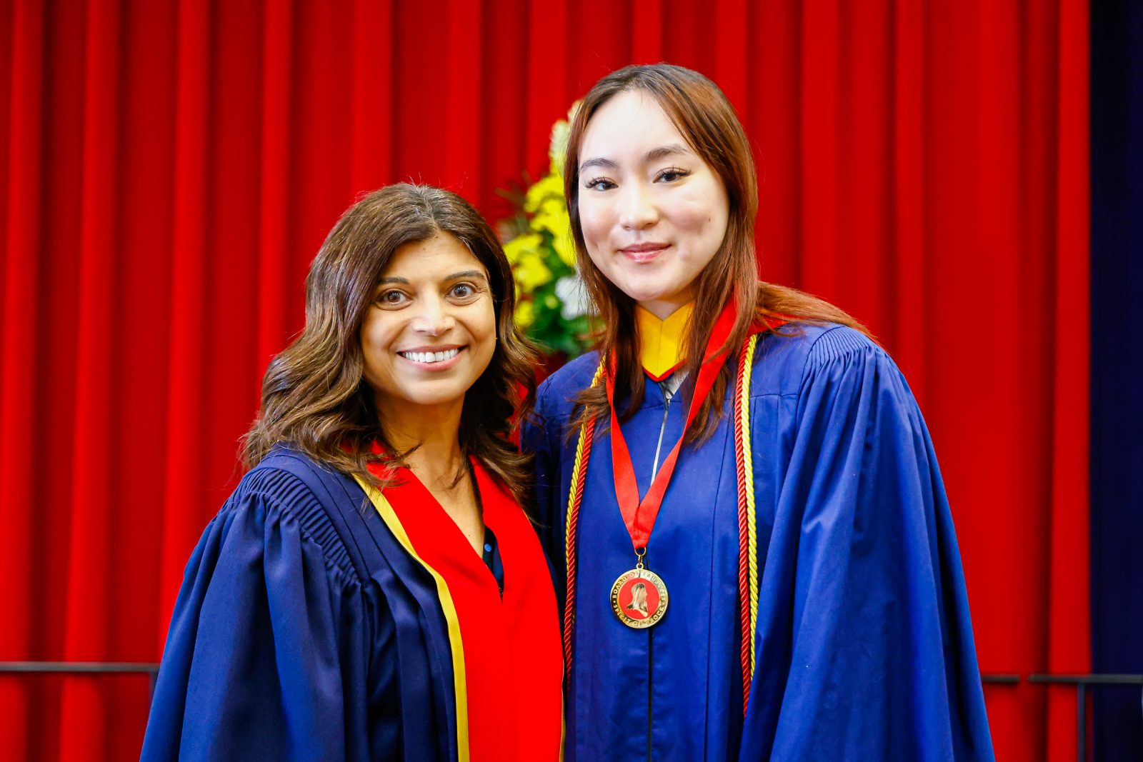 Arti Freeman (left) presents Silvana Nguyen with a Spirit of Brock medal on stage during Brock University’s Convocation ceremony. They are each wearing academic regalia.