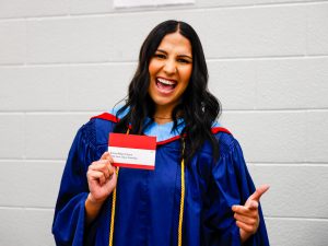 A woman in a convocation robe holds a name tag while standing in front of a white brick wall.