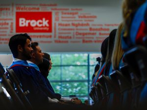 A row of people wearing convocation robes sit in a gymnasium in front of a large sign that says 'Brock University' and is surrounded by the word 'welcome' in numerous languages.