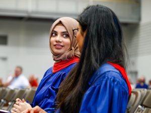 Two women in convocation robes sit in a gymnasium.