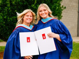 Two women in convocation robes stand outdoors next to a tree while holding their university degrees.
