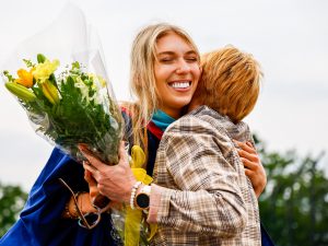 A woman in a convocation robe hugs another woman who is handing her flowers.