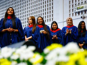 A group of women in convocation robes clap their hands while standing in a gymnasium.