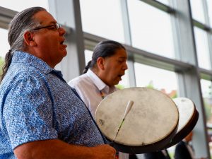 Two men play traditional drums in an indoor convocation ceremony while standing next to large windows.