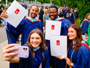 A group of students take a selfie photo while wearing their ceremonial convocation robes and holding their newly acquired university degrees.