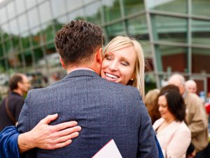 A woman and man hug while standing in a crowd outside of a large glass building.