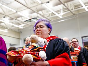A woman in a ceremonial convocation robe holds a teddy bear dressed in traditional orange and red Indigenous clothing while sitting in an indoor gymnasium.