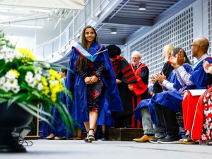 A woman walks across a stage in a robe in front of a gallery of seated people at a convocation ceremony at Brock University.
