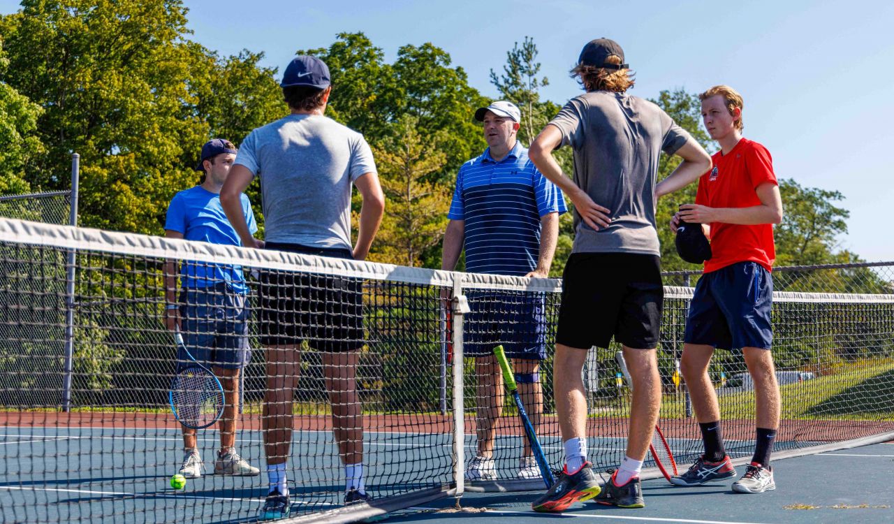 Todd Green stands behind a tennis net while four male tennis players with rackets listen to what he is saying.
