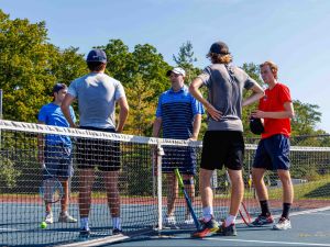 Todd Green stands behind a tennis net while four male tennis players with rackets listen to what he is saying.
