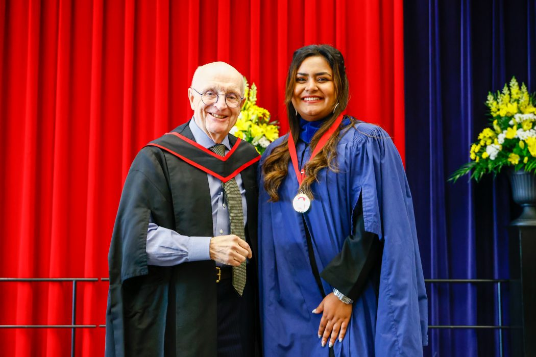 A man in academic regalia stands beside a graduate on stage during Convocation. The new graduate is wearing a Spirit of Brock medal.