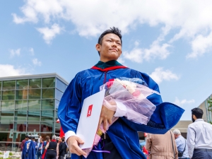 A Brock University graduate poses in his graduation gown while holding a diploma and flowers.