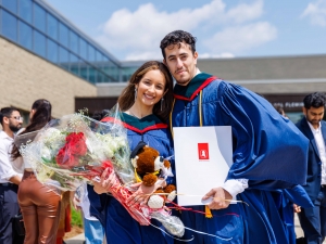 Two Brock University graduates in Convocation gowns pose with a diploma and bouquets of flowers.