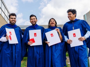Four Brock University graduates stand side-by-side in their graduation gowns holding their degrees while smiling for a photo.