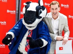 A new graduate poses in front of a backdrop with a badger mascot at graduation.