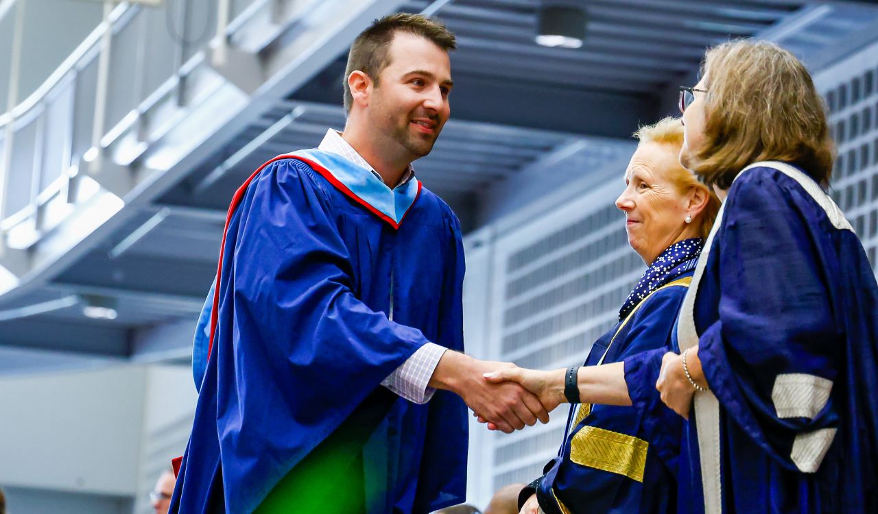 A person in a blue Convocation gown shakes the hands a woman while crossing a graduation stage.