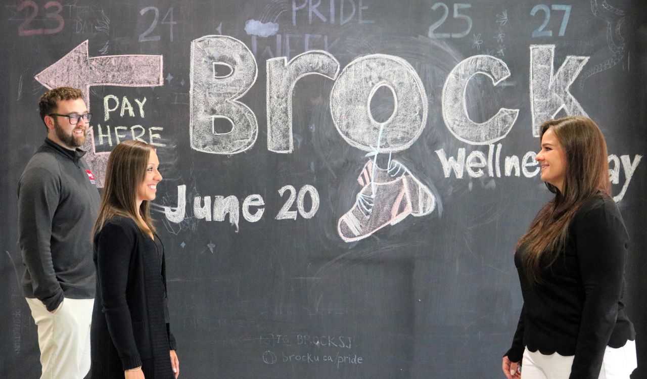 Two people walk toward another person standing in front of a chalkboard wall sign promoting Brock’s Employee Wellness Day.