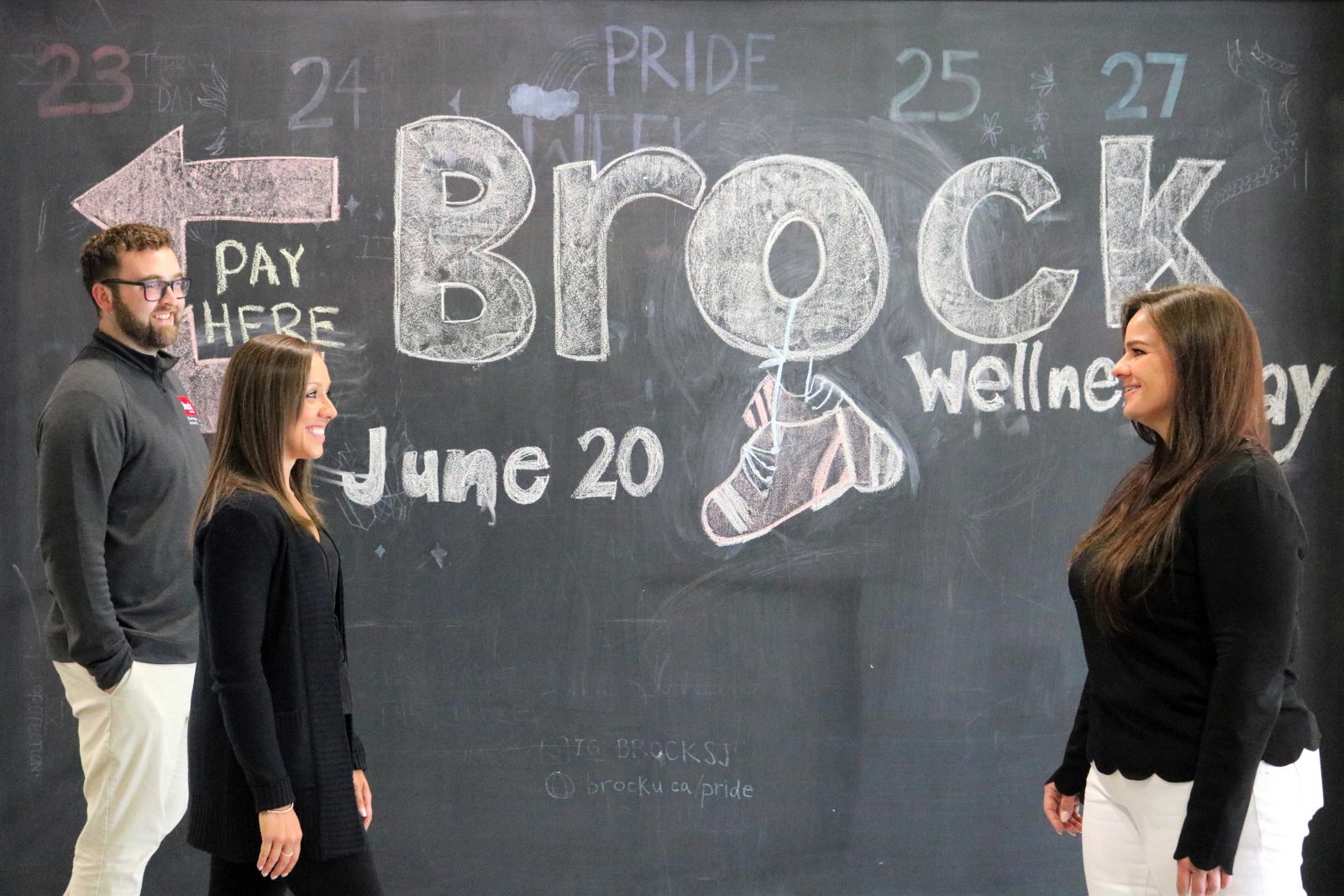 Two people walk toward another person standing in front of a chalkboard wall sign promoting Brock’s Employee Wellness Day.