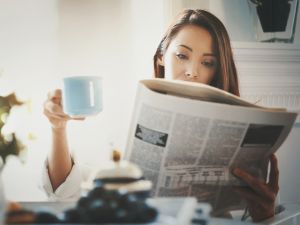 A woman sits holding a coffee cup in one hand and a newspaper in the other.