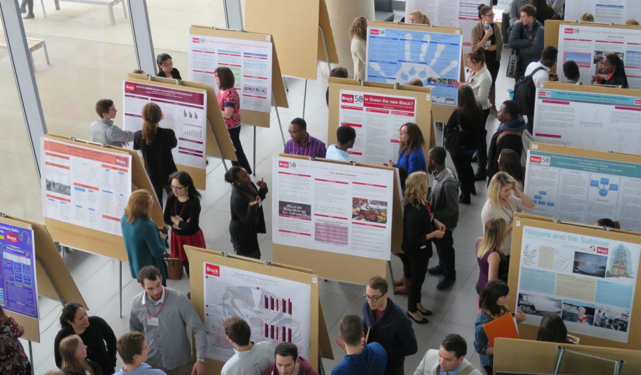 A large crowd of people look at more than a dozen research poster presentations, which are displayed on cork board easels and lined in rows in a large and brightly lit hallway at Brock University.
