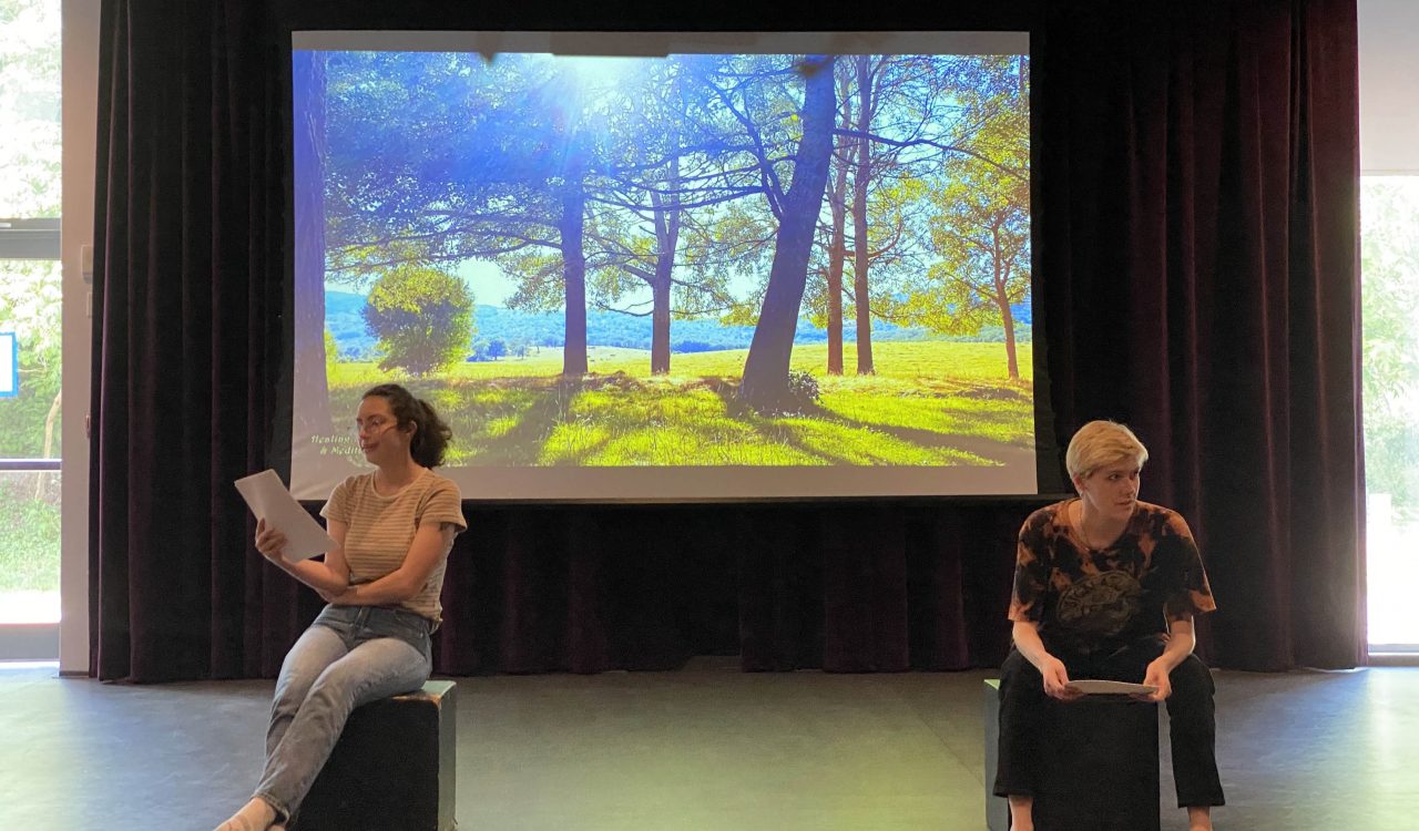 Two students in street clothing sit in a dramatic arts studio with a backdrop image of two large trees on a sunny day behind them. The students are engaged with a script during a workshop session.