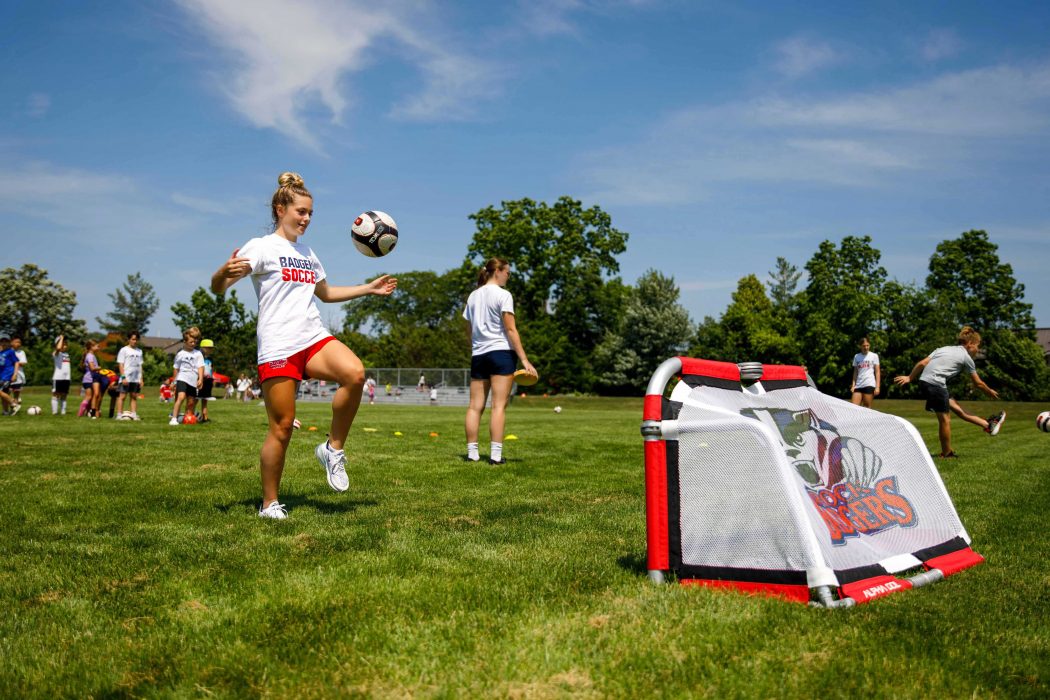 A woman juggles a soccer ball during a kids sports camp outdoors on a green field under blue skies.