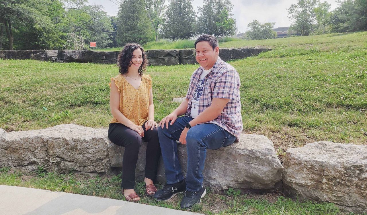 A woman and man sit next to each other on a rock in front of a grassy field.