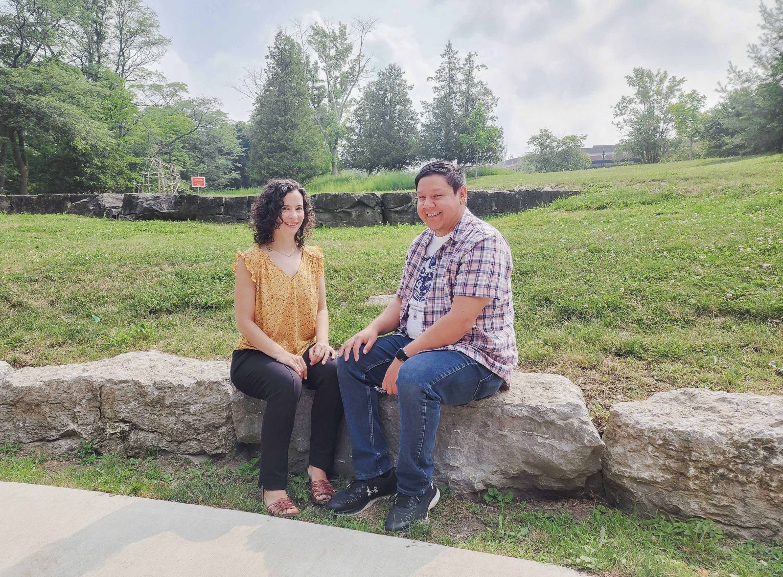 A woman and man sit next to each other on a rock in front of a grassy field.
