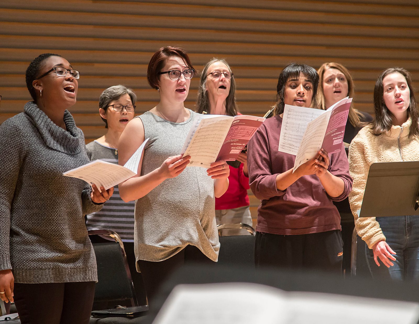 A group of Brock University choir members sing on stage holding sheet music and looking in the direction of the conductors.