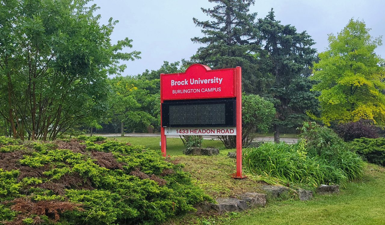 An outdoor red sign reads 'Brock University Burlington Campus.' It is set against trees and green landscape with rocks along a pathway. The sky is overcast.
