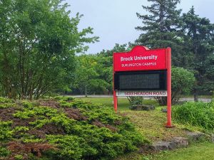 An outdoor red sign reads 'Brock University Burlington Campus.' It is set against trees and green landscape with rocks along a pathway. The sky is overcast.
