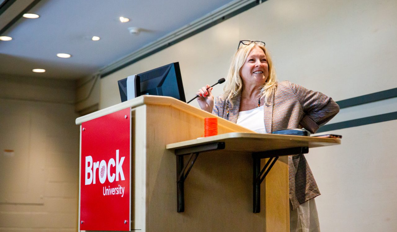 Cindy Biancaniello, Director of Brock's Hadiya’dagénhahs First Nations, Métis and Inuit Student Centre, stands at a podium speaking into a microphone.