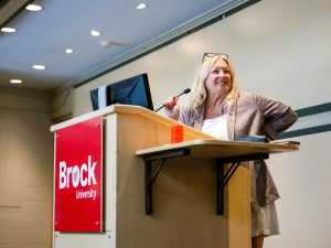 Cindy Biancaniello, Director of Brock's Hadiya’dagénhahs First Nations, Métis and Inuit Student Centre, stands at a podium speaking into a microphone.