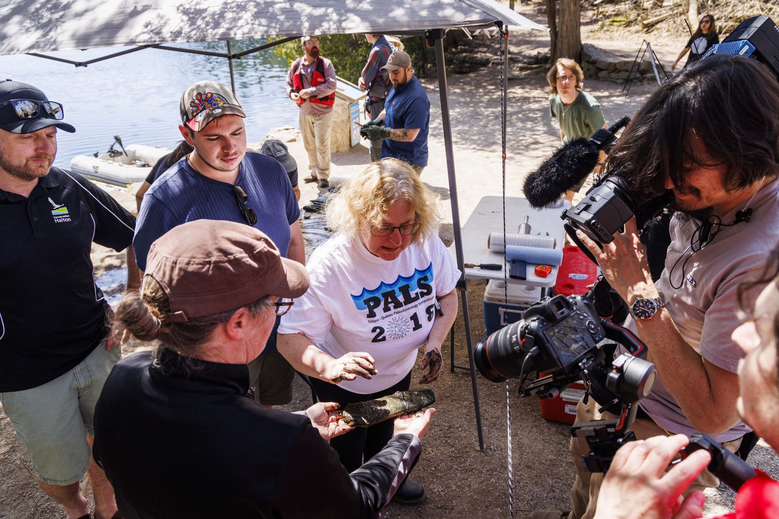 University Professor of Earth Sciences Francine McCarthy (centre) points to a slab being held by a woman wering a cap and black shirt, with two cameras pointing at them while members of the research team look on.