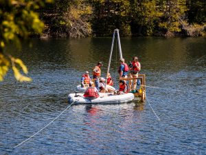 Three men standing, and one woman sitting, on a raft with poles that resemble a tripod sticking up, with two inflatable dinghies carrying a total of four people sit next to the raft.