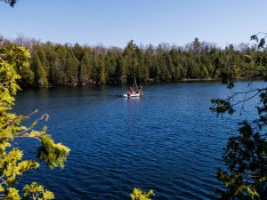 A long-distance photo of dark-blue Crawford Lake ringed by a forest of mostly coniferous trees. At the centre of the lake is a platform with two steel poles sticking up and three people standing on the platform wearing red and orange lifejackets. A white inflatable boat with one person sitting in it wearing a red life jacket sits in front of the raft.