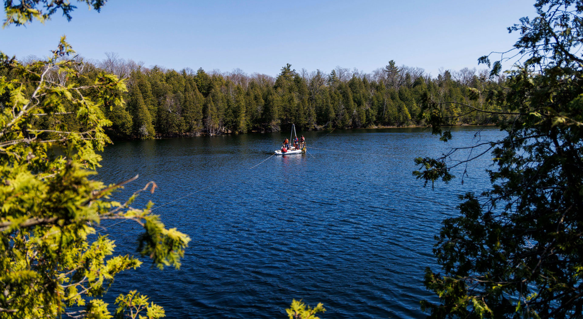 A long-distance photo of dark-blue Crawford Lake ringed by a forest of mostly coniferous trees. At the centre of the lake is a platform with two steel poles sticking up and three people standing on the platform wearing red and orange lifejackets. A white inflatable boat with one person sitting in it wearing a red life jacket sits in front of the raft.