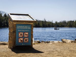 A short stone pillar, with a sign that reads “Crawford Lake is a rare, meromictic lake,” stands in front of Crawford Lake and trees in the background.