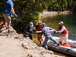 Then-Carleton undergraduate student Krysten Lafond (now at Queen's University), Tim Patterson (Professor of Earth Sciences, Carleton University), Paul Hamilton (Research & Collections, Canadian Museum of Nature) and Maxim Ralchenko (Carleton University) carry a freeze core resembling a chocolate-brown slab from an inflatable dingy to the shoreline.