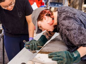 University students Anne Nguyen (left) and then-Carleton undergraduate student Krysten Lafond (now at Queen's University) are hunched over a dark-brown slab laying on a table, with Lafond holding a chisel.