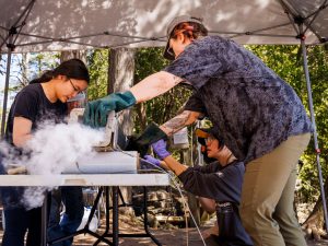 Carleton University students (left to right) Emily Shi, Anne Nguyen and then-Carleton undergraduate student Krysten Lafond (now at Queen's University bend over a tube on a table, chiseling frozen mud off the tube, with steam coming out of an opening in the tube.