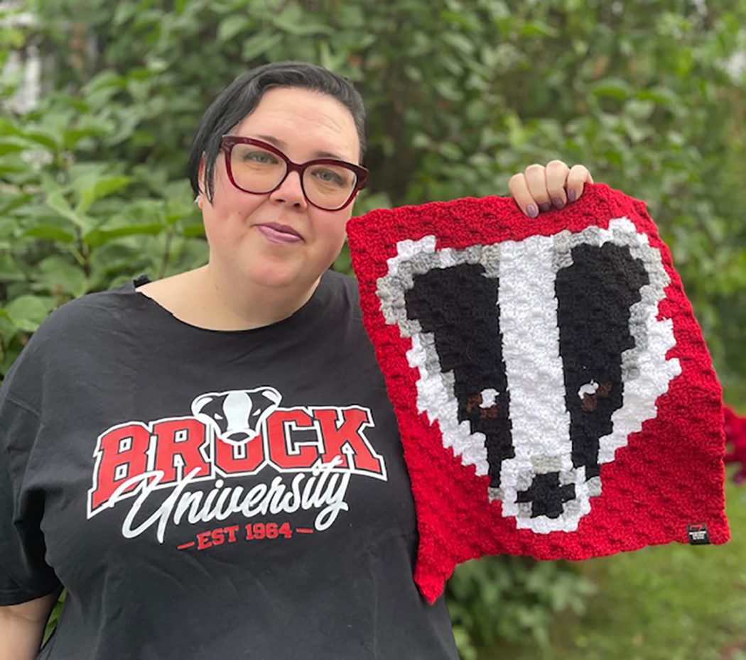 Emily Hammond holds up a crochet design she made of Brock University’s Badger mascot while wearing a T-shirt with the image that inspired her creation.