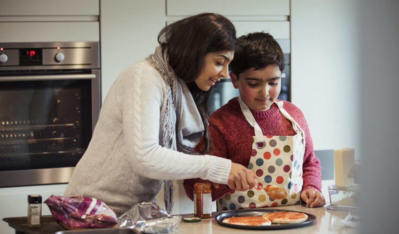 A woman wearing a white sweater leans towards her son wearing a red sweater and polka-dotted apron as they both hold a spoon covered in tomato sauce that they are spreading over mini-pizzas on the kitchen counter.