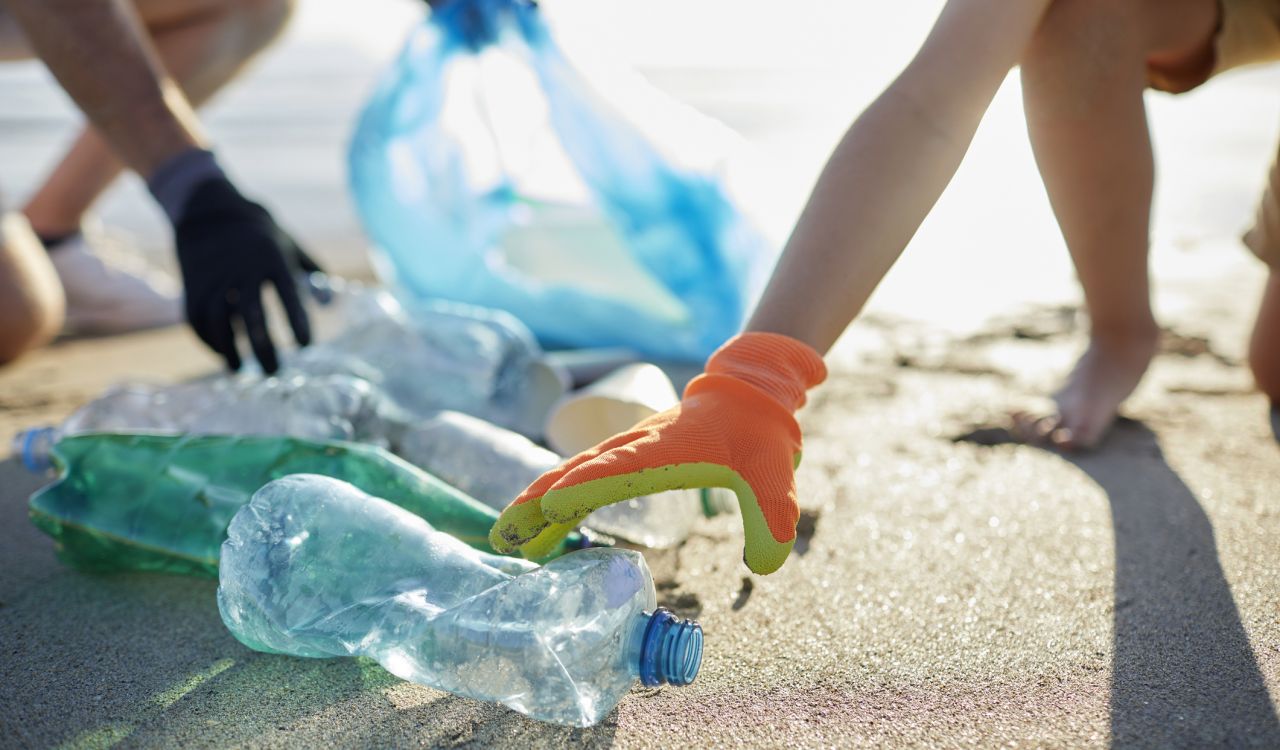 Close-up of arms and hands wearing gloves reaching for a group of plastic bottles laying on a beach, with a blue recycling bag in the background.